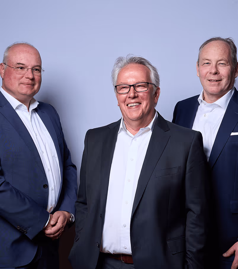 Drei Geschäftsleute in formeller Kleidung, die nebeneinander stehen und mit selbstbewussten Gesichtsausdrücken in die Kamera blicken. Der Hintergrund ist eine einfache, hellgraue Wand.