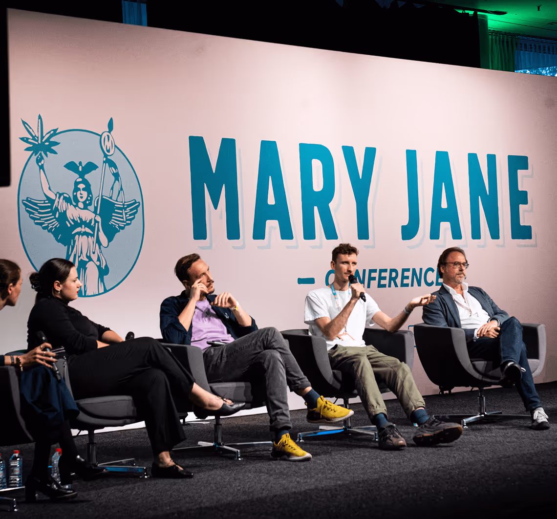 Eine Konferenzbühne mit einem Hintergrund, auf dem „MARY JANE - CONFERENCE“ steht. Eine Gruppe von fünf Personen sitzt auf der Bühne und diskutiert ein Thema. Eine Person spricht in ein Mikrofon, während die anderen aufmerksam zuhören.