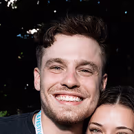 Eine Nahaufnahme eines Mannes, der breit lächelt und ein schwarzes T-Shirt trägt. Um seinen Hals hängt ein Lanyard. Der Hintergrund ist draußen mit natürlichem Licht.