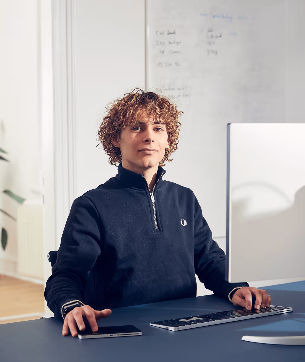 Ein gutaussehender Mann sitzt an einem Schreibtisch im Büro, schaut in die Kamera und hat einen neutralen Gesichtsausdruck. Er trägt einen blauen Zip-Pullover und ist von modernen Bürogeräten wie einer Tastatur, einem Tablet und einem Handy umgeben.