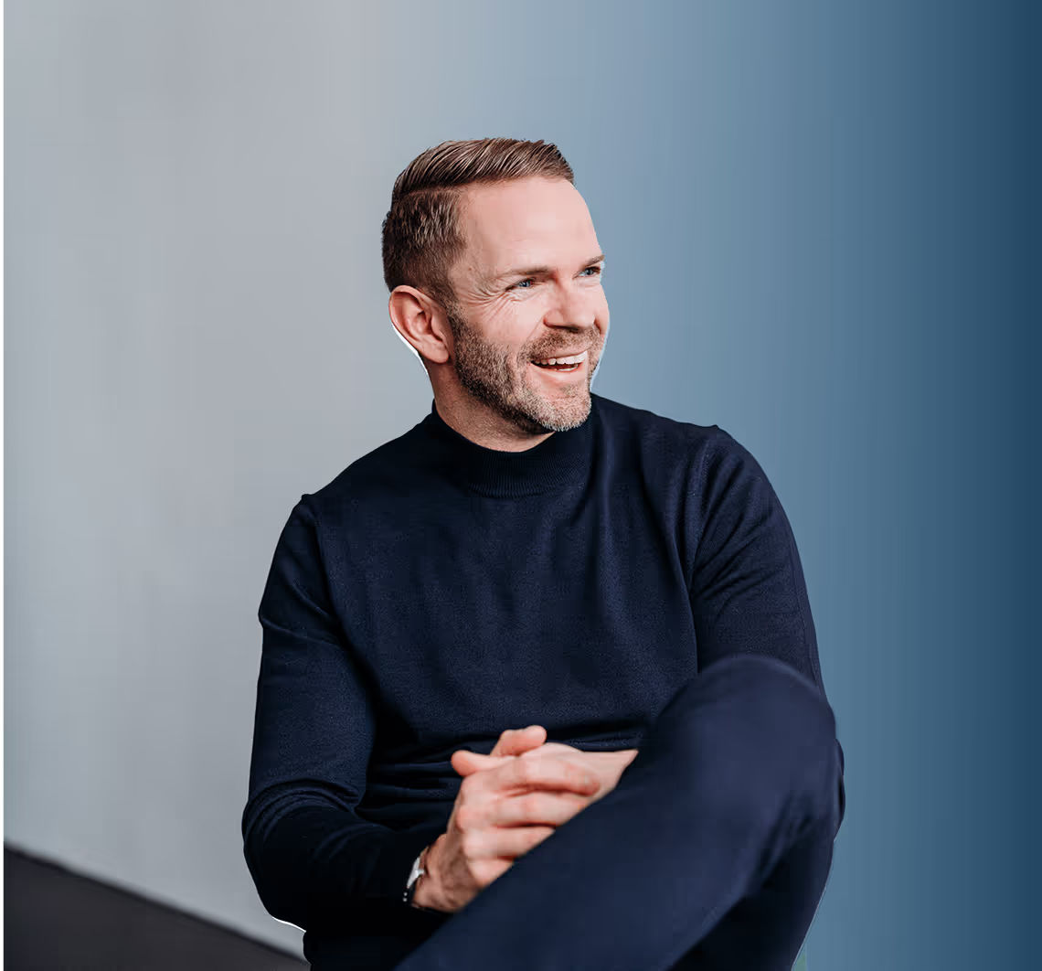 Smiling man with short hair and beard wearing a black sweater sitting against a gradient blue and gray background.