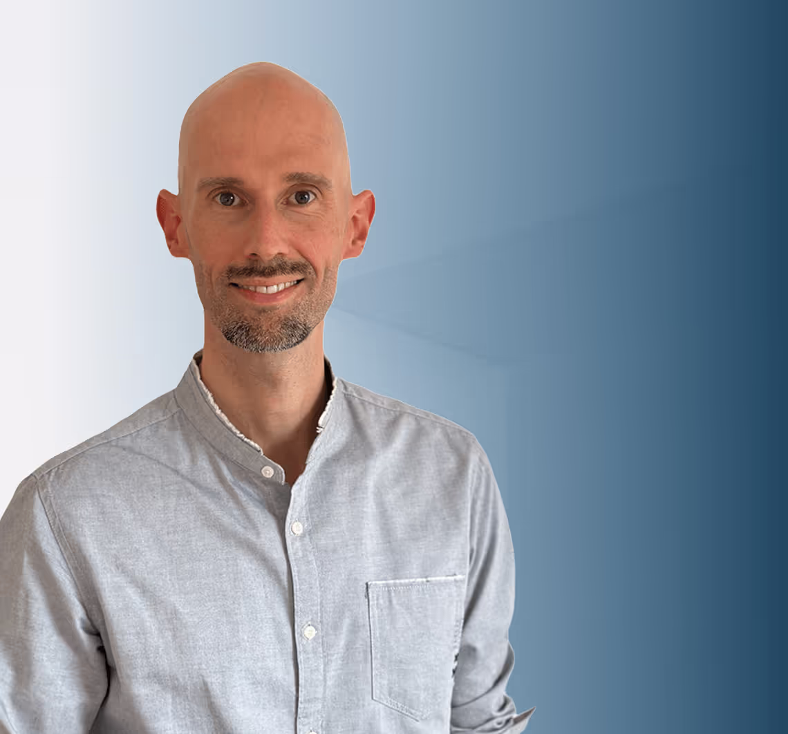 Smiling bald man with a short beard wearing a light grey button-up shirt against a blue gradient background.